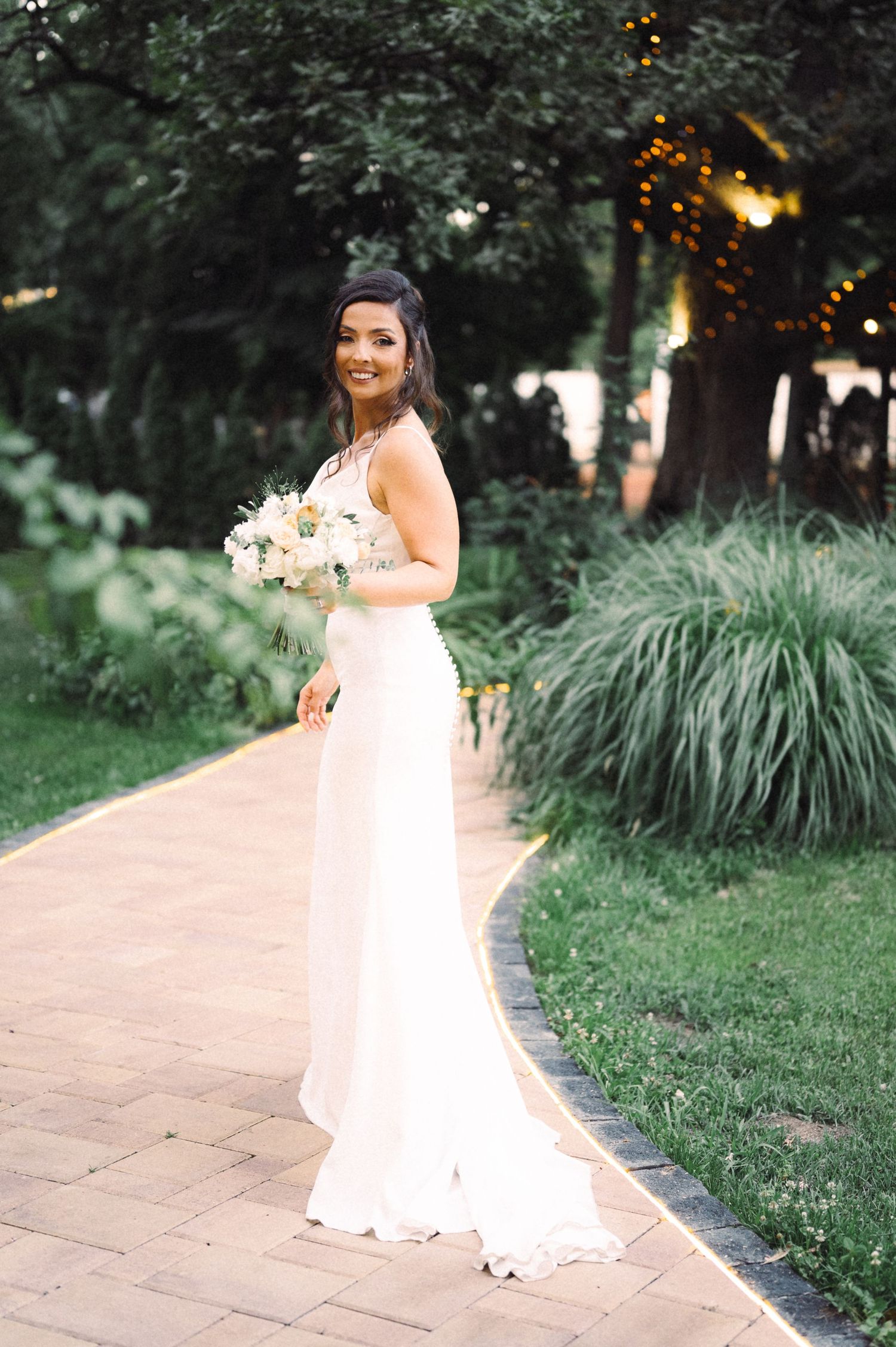 Elegant wedding dress silhouette with flowing fabric against a garden backdrop with twinkle lights.