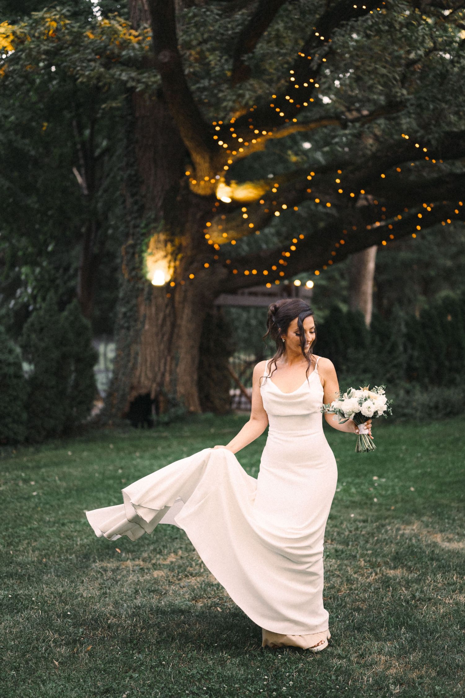 Flowing white wedding gown twirls in a garden setting with twinkling lights in the background.