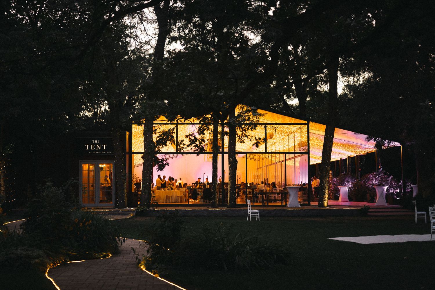 Modern glass wedding venue illuminated at dusk with trees silhouetted against sunset sky.