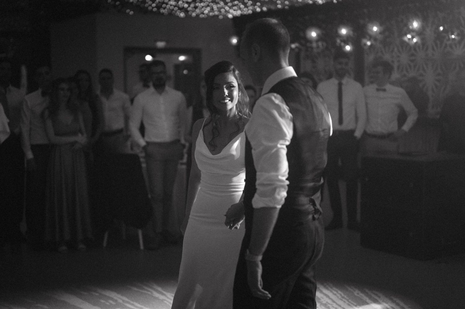 A black and white photo of a couple's first dance at a wedding reception with guests watching in the background.
