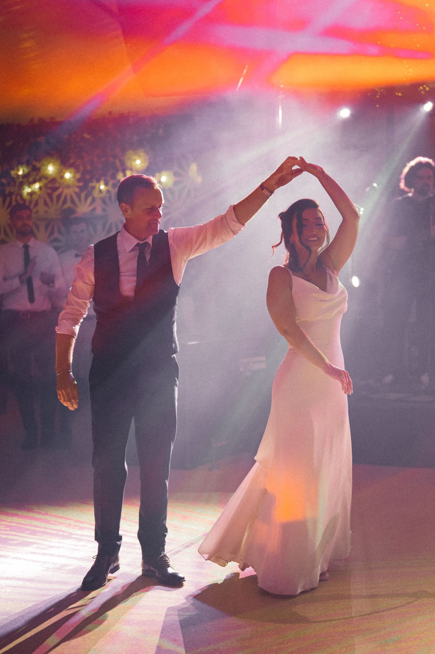 A couple performs a romantic twirl during their wedding dance under colorful mood lighting.