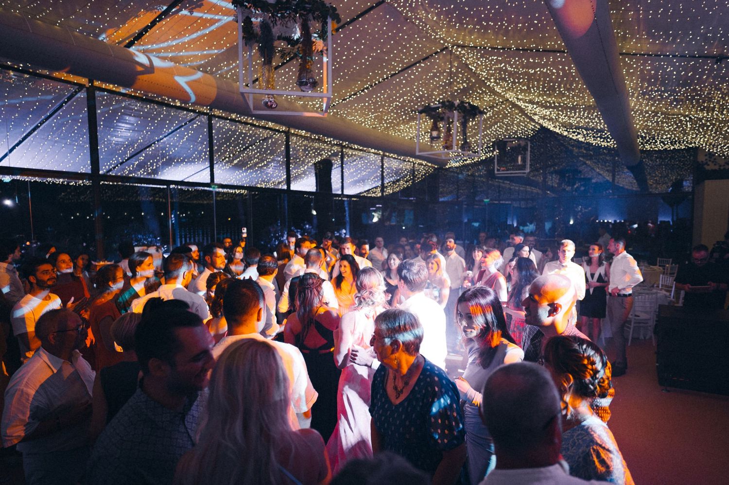 A crowded wedding reception dance floor illuminated by colorful lights and string lighting overhead.