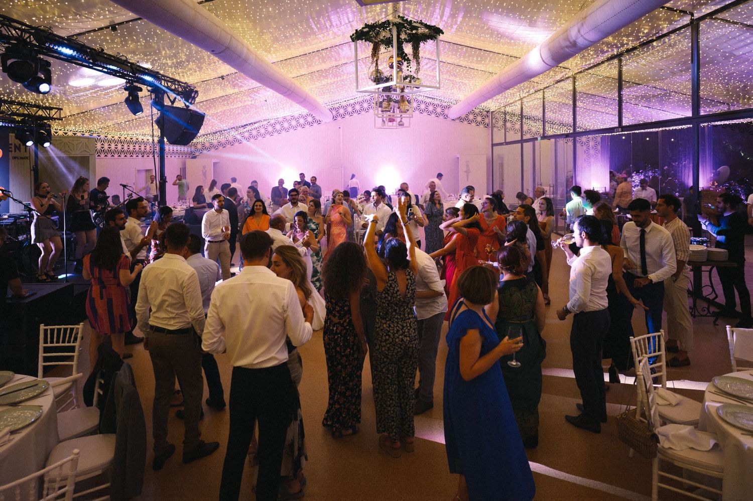 Large group dancing at an indoor reception venue with purple lighting and white chairs.