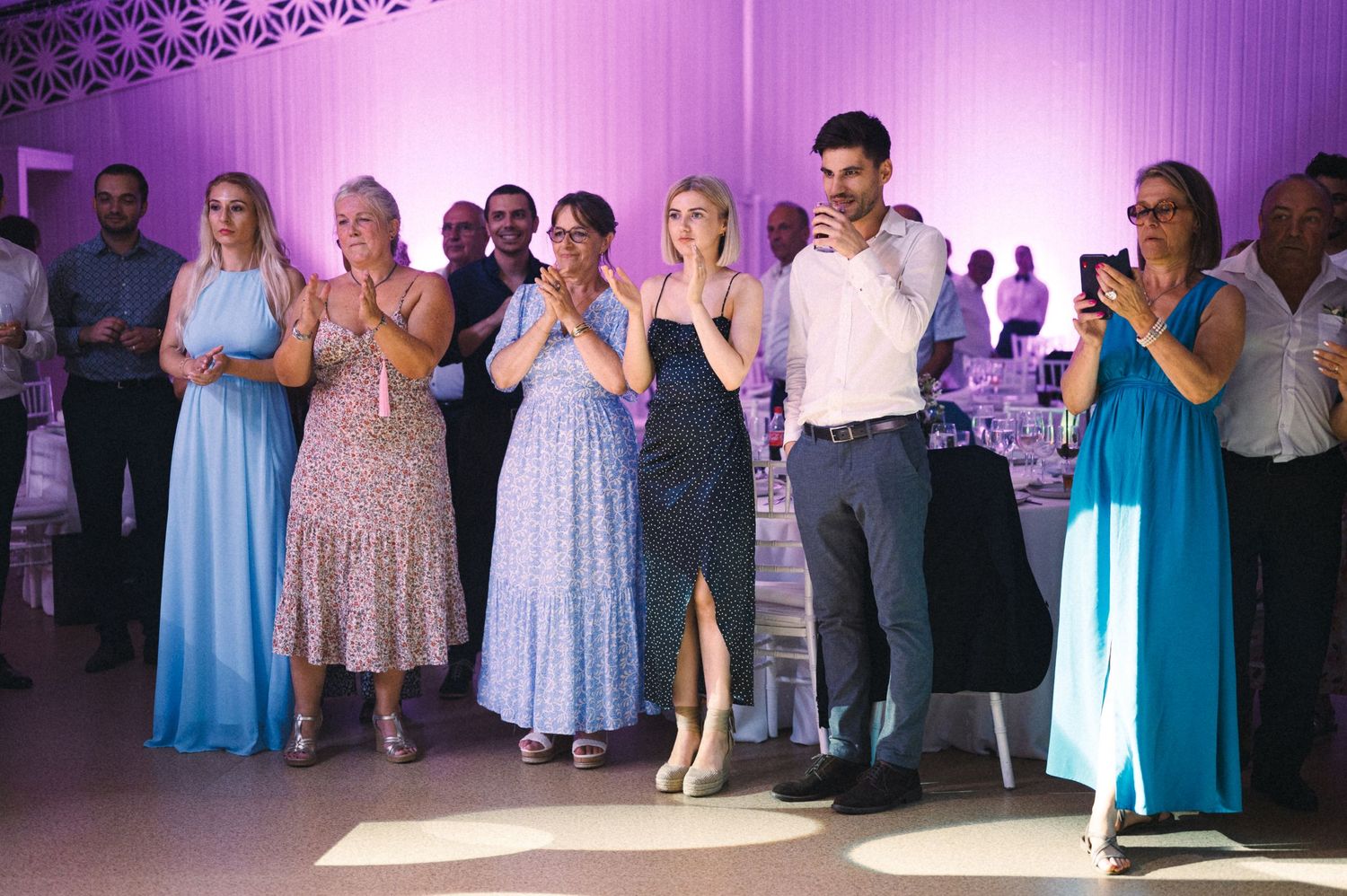 Wedding guests in formal attire standing in line at reception with purple ambient lighting.