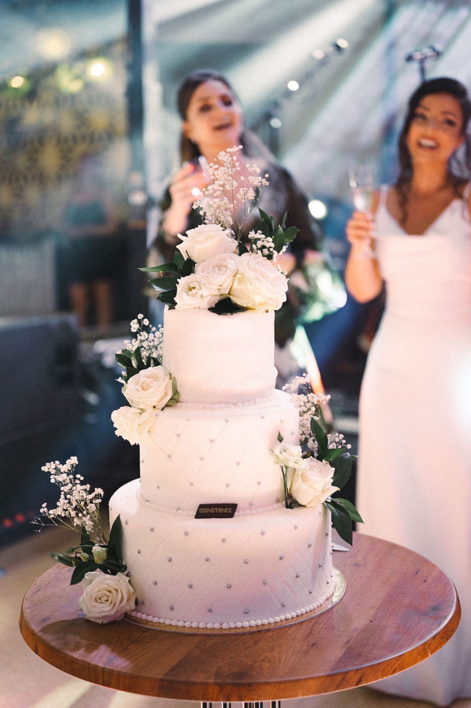 Elegant white wedding cake decorated with roses and silver accents on wooden stand.