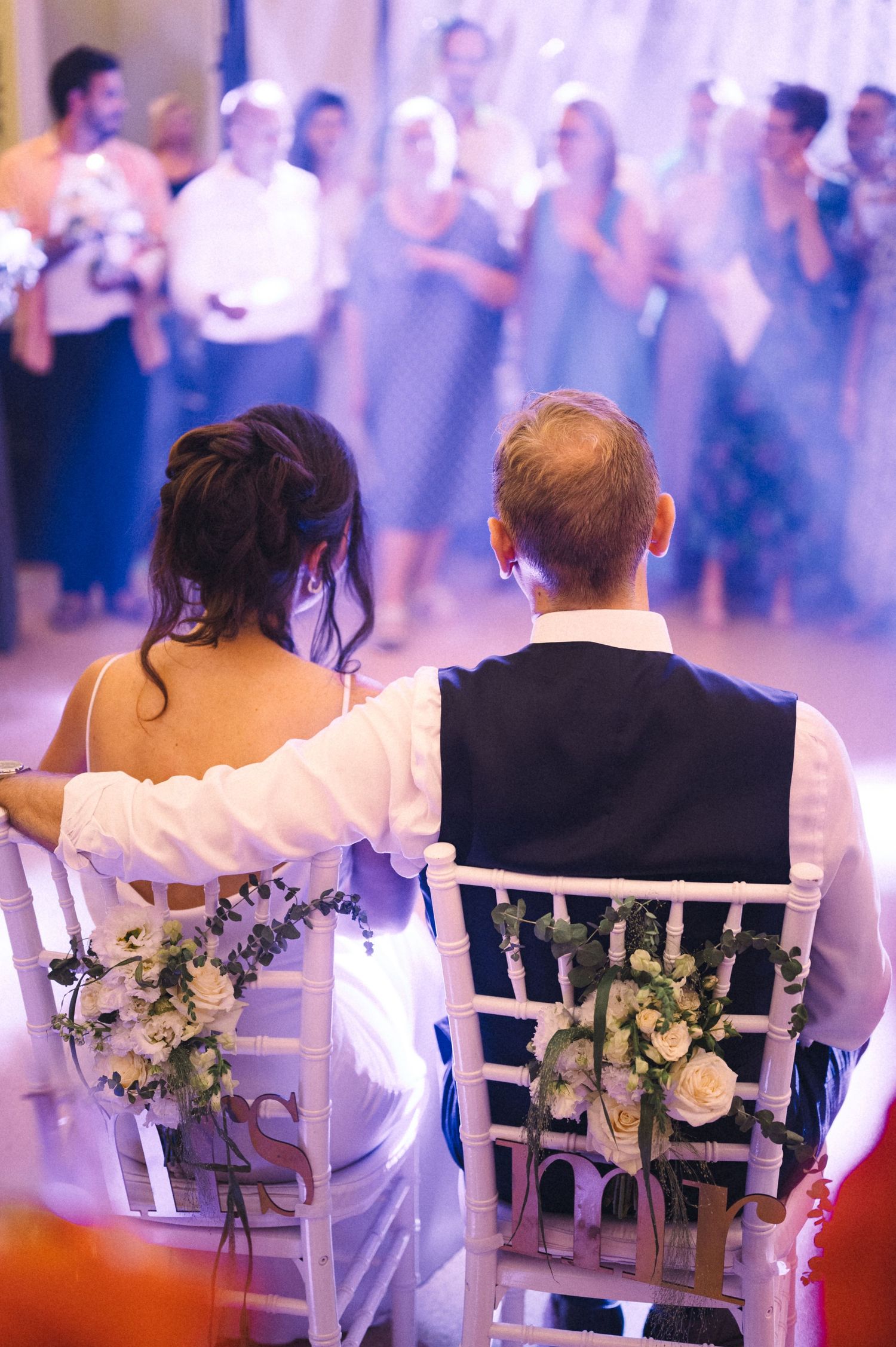 Newlyweds sitting in decorated chairs with floral arrangements during their wedding reception.