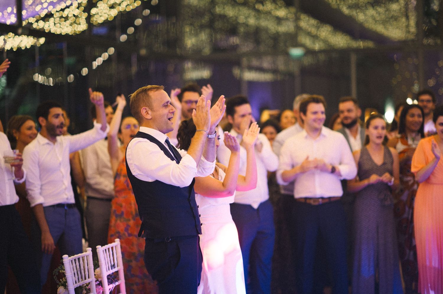 Guests celebrate and applaud during a wedding reception under twinkle lights.