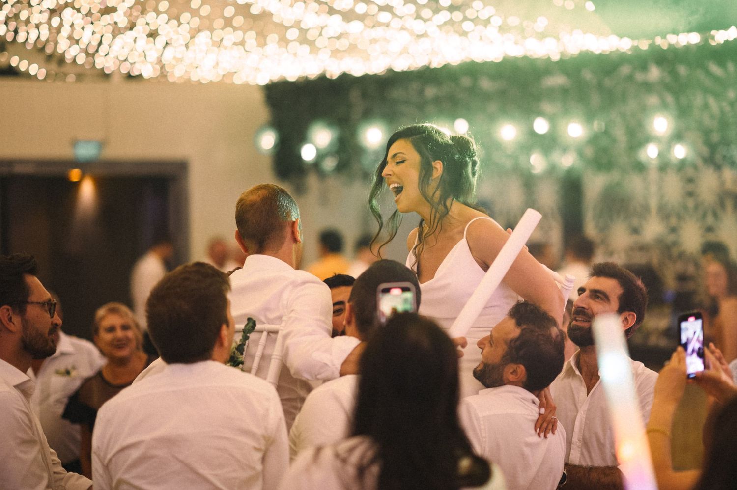 Wedding guests lift the celebrants during a lively dance party under twinkling lights at a reception.