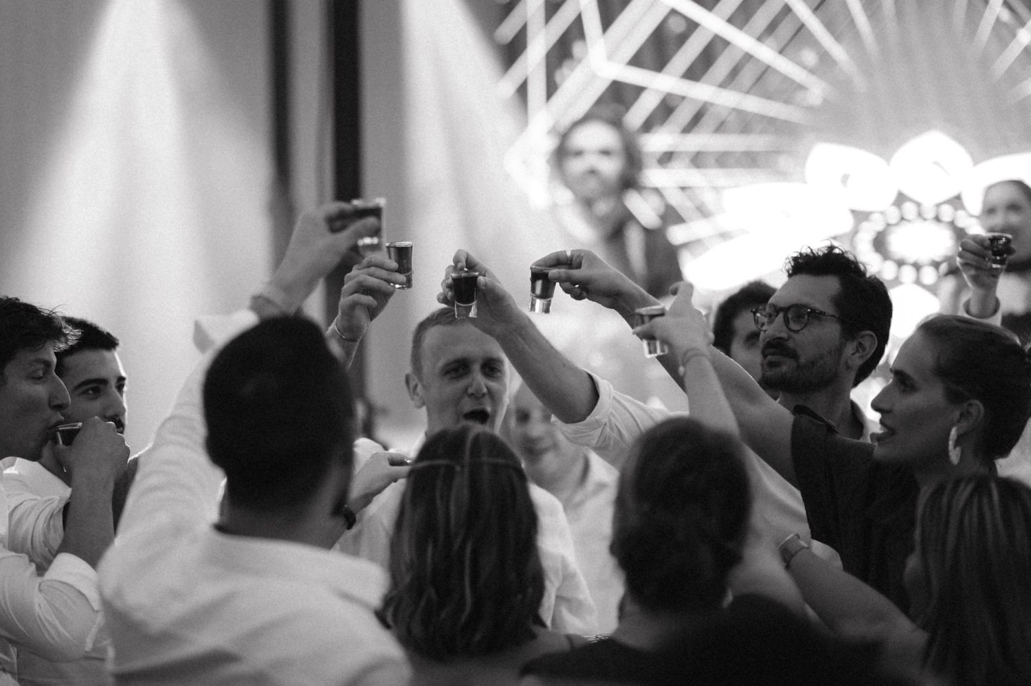 Black and white shot of party guests raising glasses for a celebration toast under bright spotlights.
