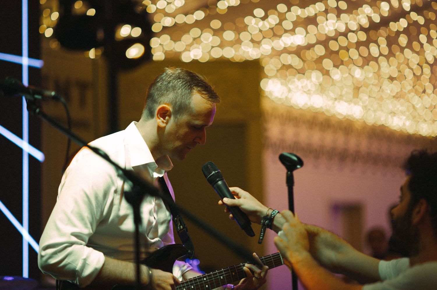 Wedding band performer plays guitar under warm string lights during evening reception.