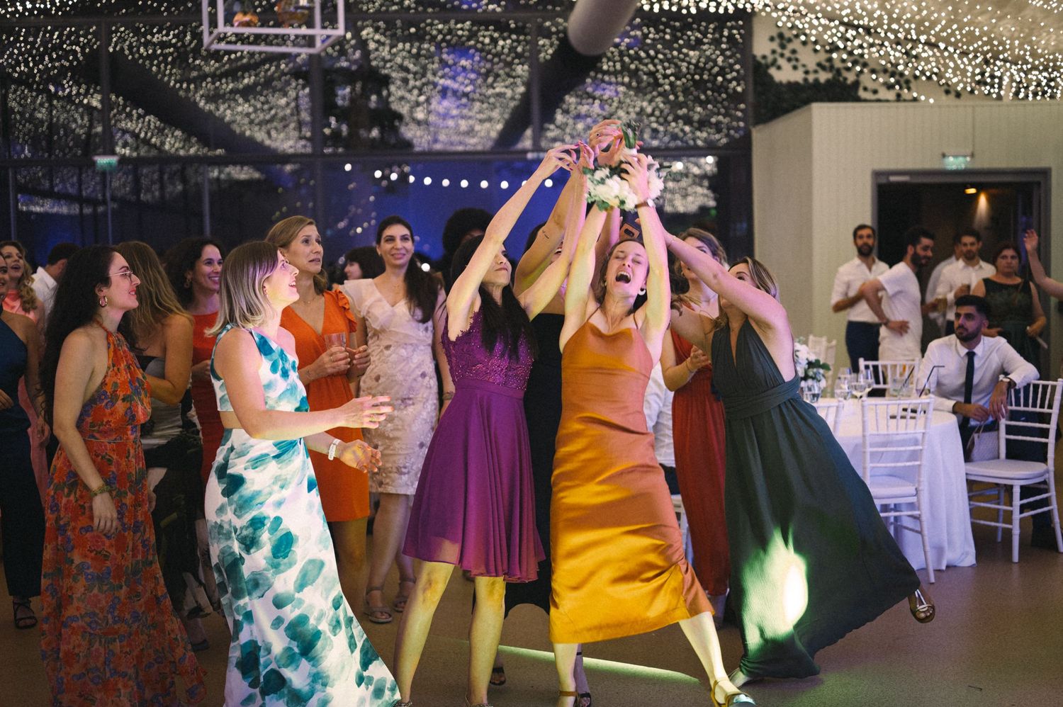 Wedding reception guests in vibrant dresses reaching for bouquet toss under string lights.