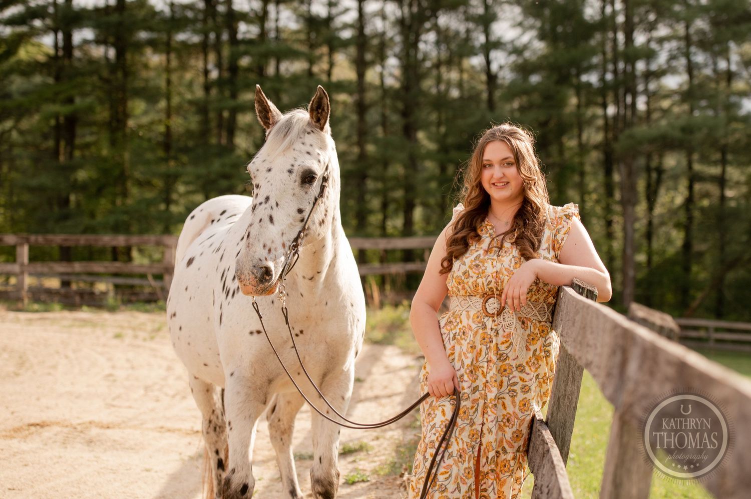 A spotted white horse and companion share a moment in an outdoor arena surrounded by pine trees.