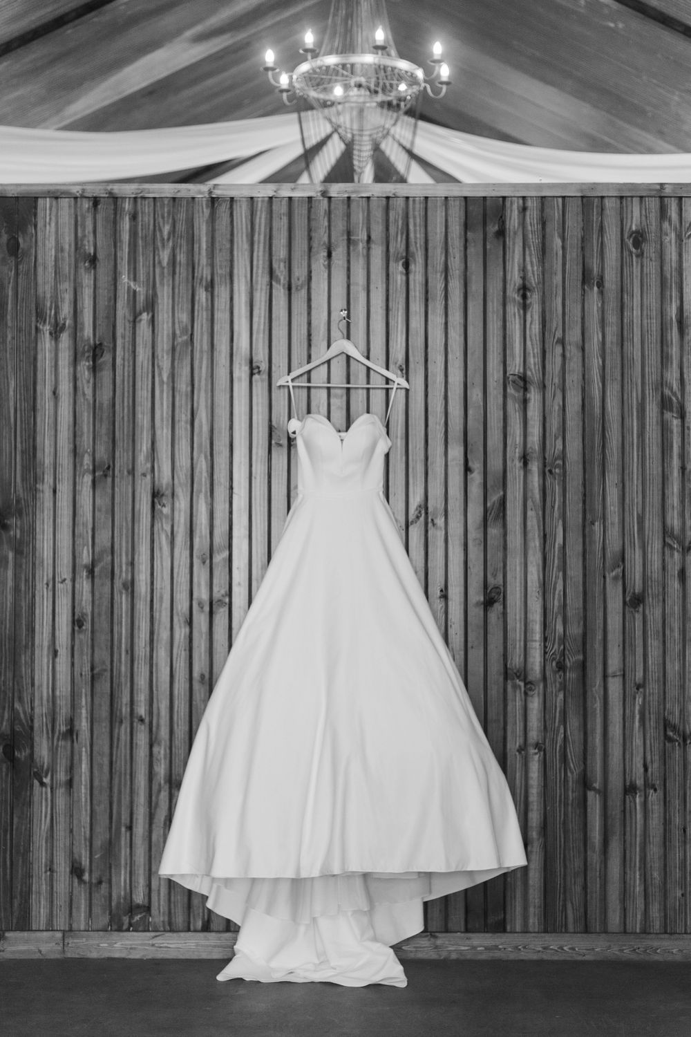 A classic white wedding dress hangs against rustic wooden walls under a chandelier.