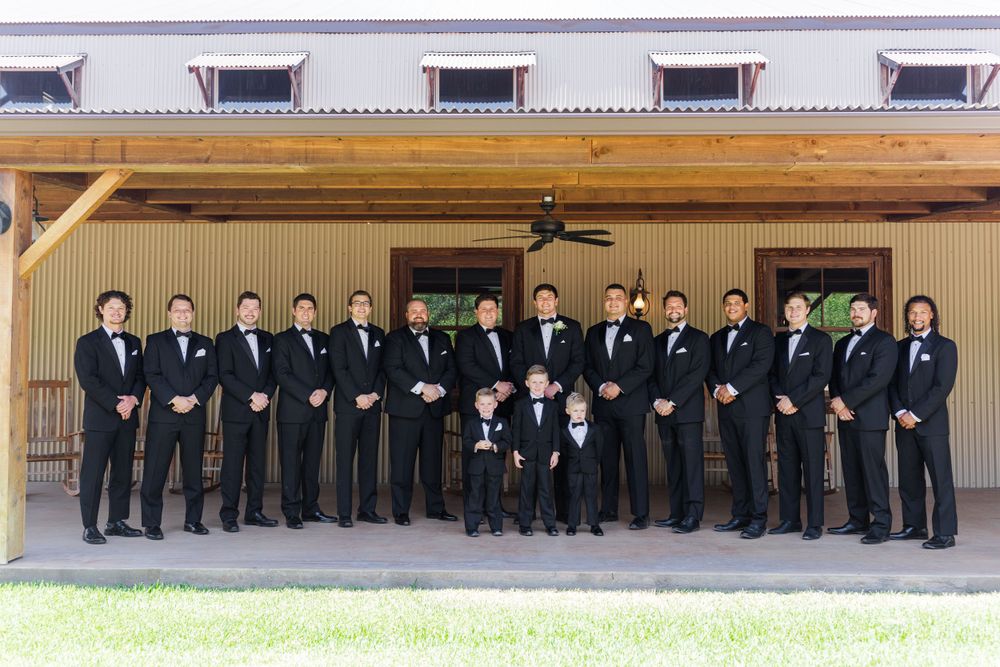 Formal group portrait of groomsmen and ring bearers in black tuxedos under covered porch.