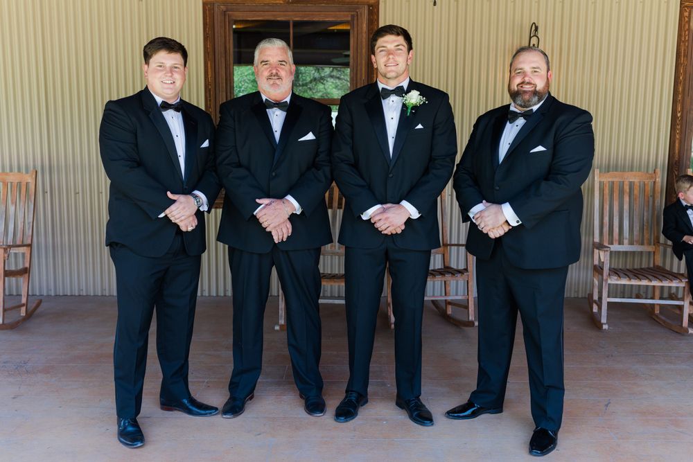 Four groomsmen in classic black tuxedos stand formally on a porch.