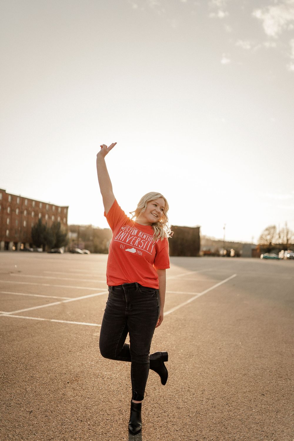 Joyful pose with one leg raised in an empty parking lot at sunset wearing orange shirt and black jeans.
