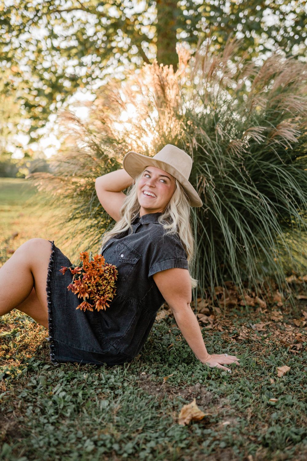 Woman in casual dress and hat sits beside ornamental grasses during golden hour sunset.