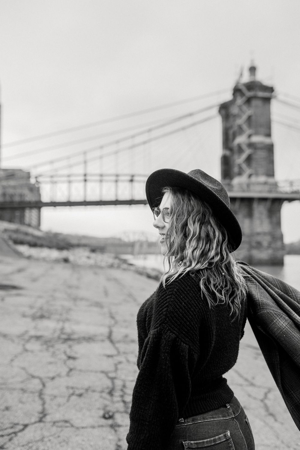 Black and white portrait near historic bridge wearing dark sweater and wide-brimmed hat.
