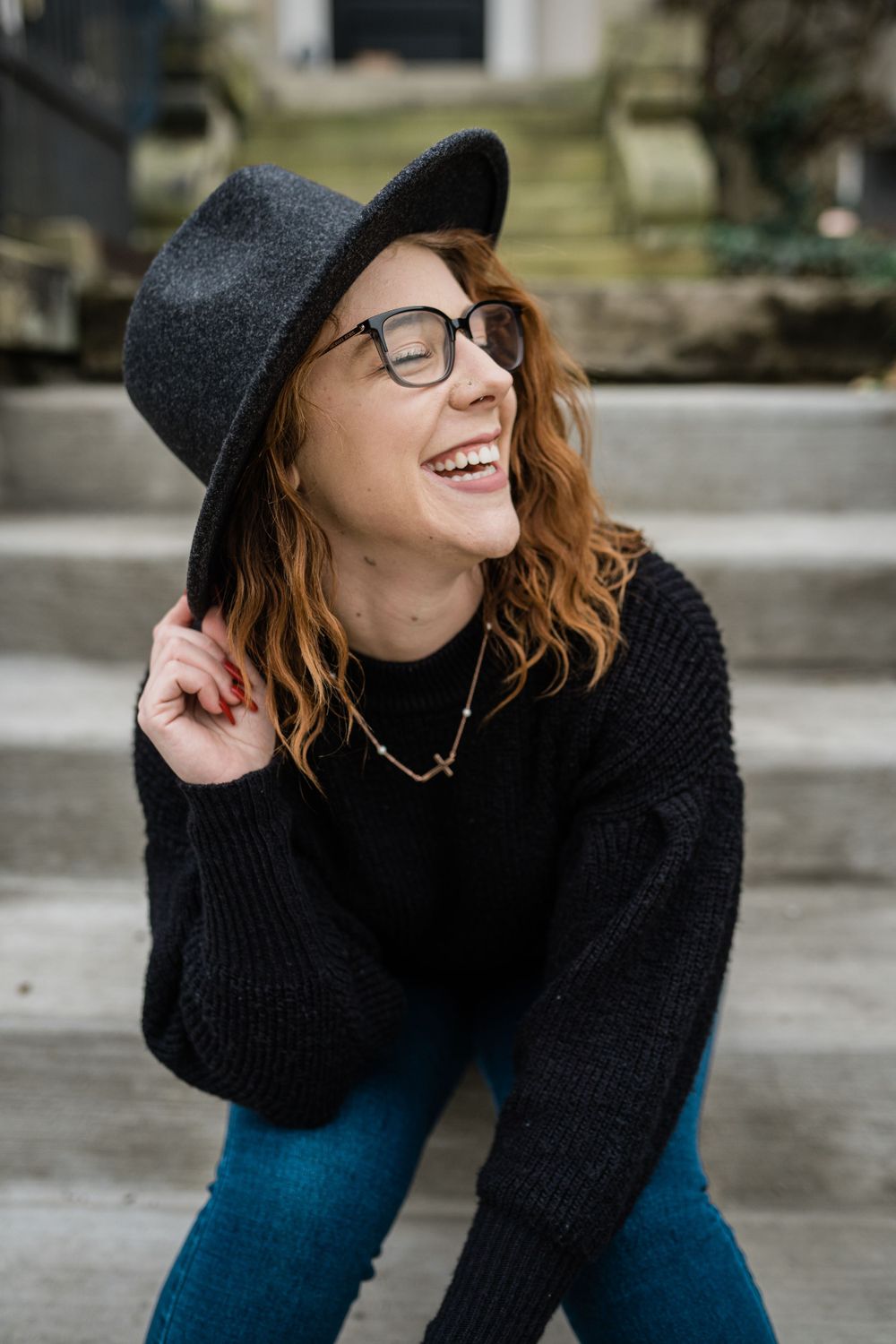 Casual portrait on steps wearing a black hat, sweater, glasses and denim with a bright genuine smile.
