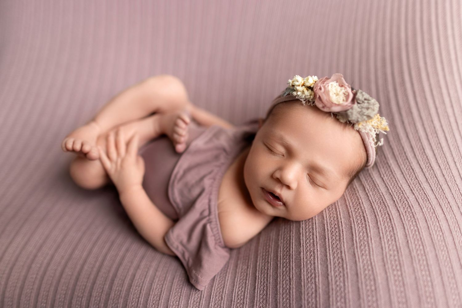 Sleeping newborn baby wrapped in purple cloth with delicate floral headband on textured pink background.