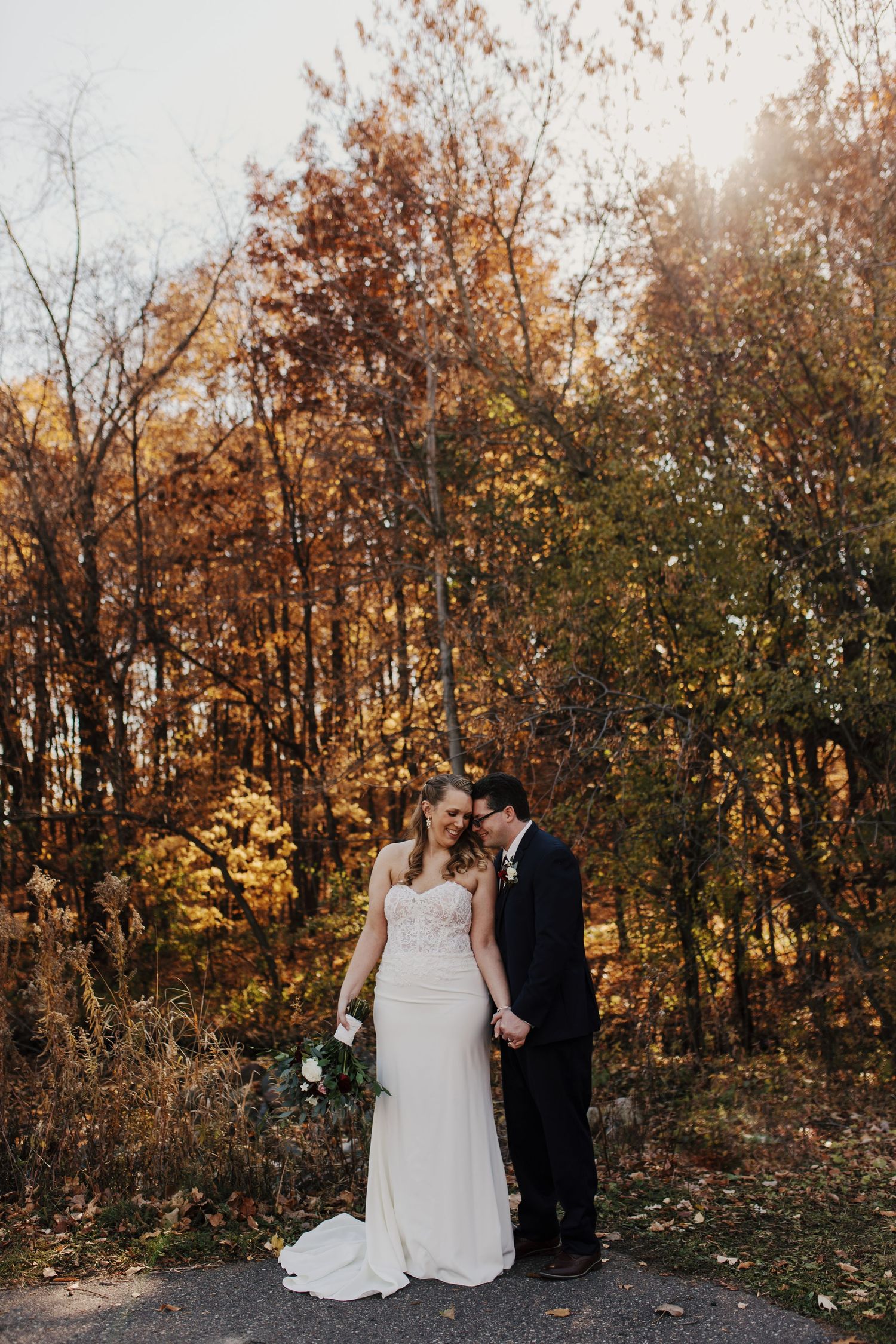 Couple holding hands while standing on path surrounded by golden autumn foliage.