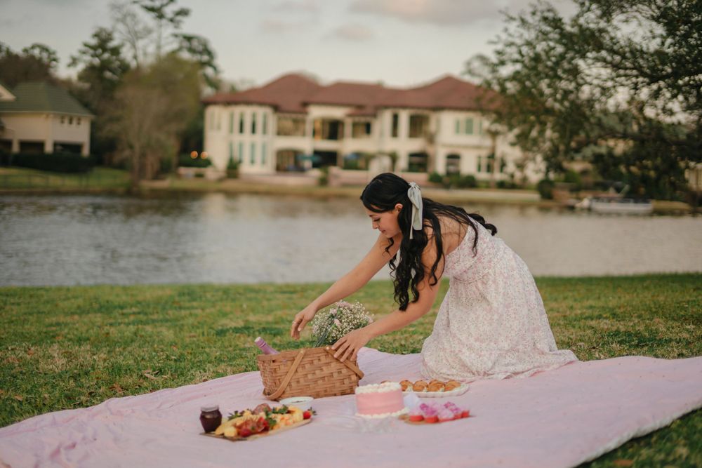 A picnic scene by a lakeside with a pink blanket and wicker basket against luxury homes in background.
