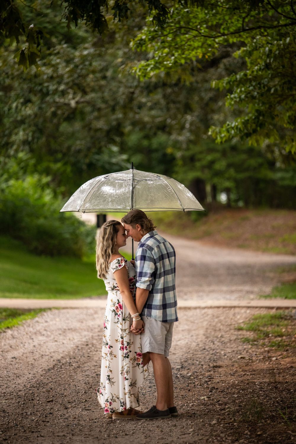 A couple shares a romantic moment under a clear umbrella on a tree-lined dirt path in a park.