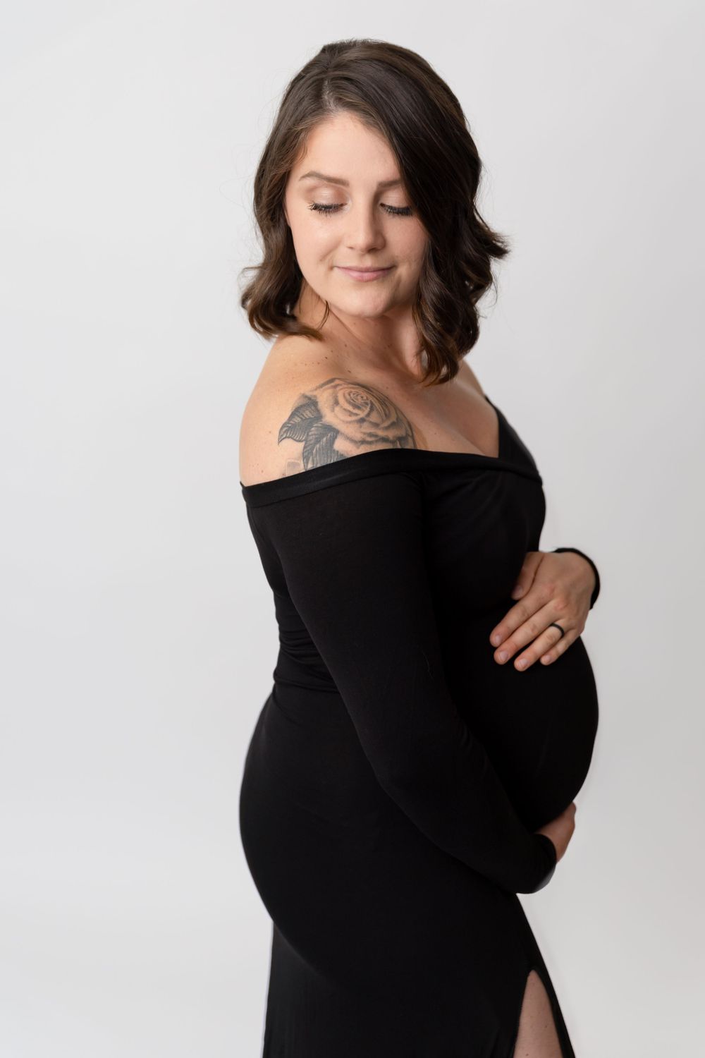 Expectant woman in elegant black off-shoulder dress poses against white backdrop showing visible shoulder tattoo.