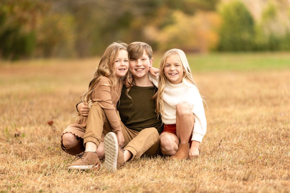 Three children sitting together in a grassy field on a sunny autumn day.