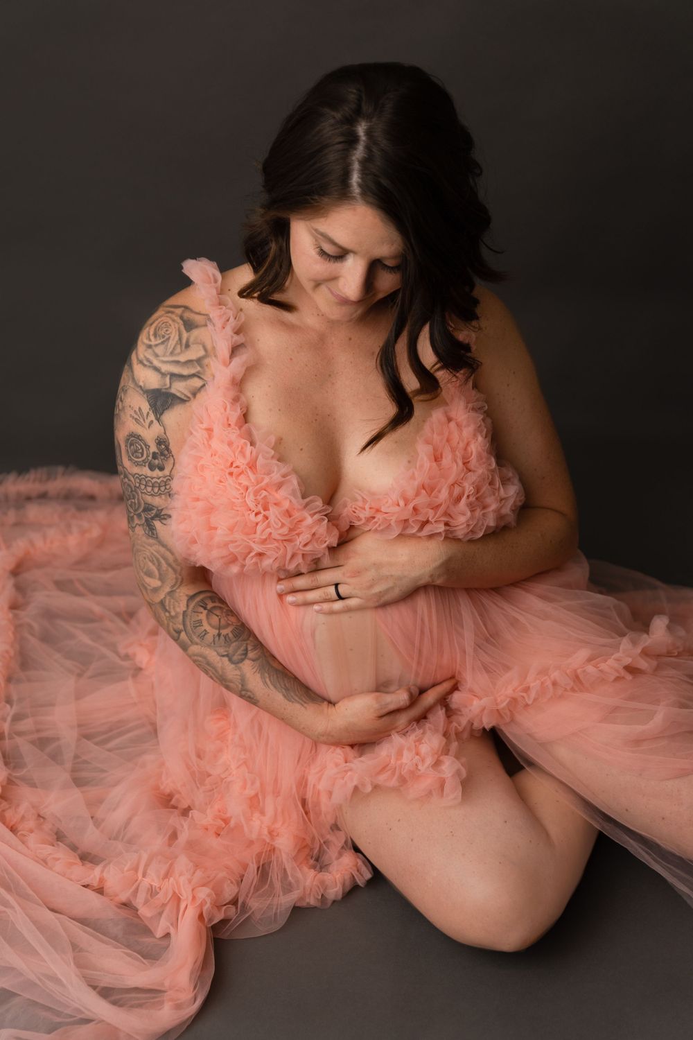 A woman in a peach tulle dress poses for a maternity photo against a dark background.