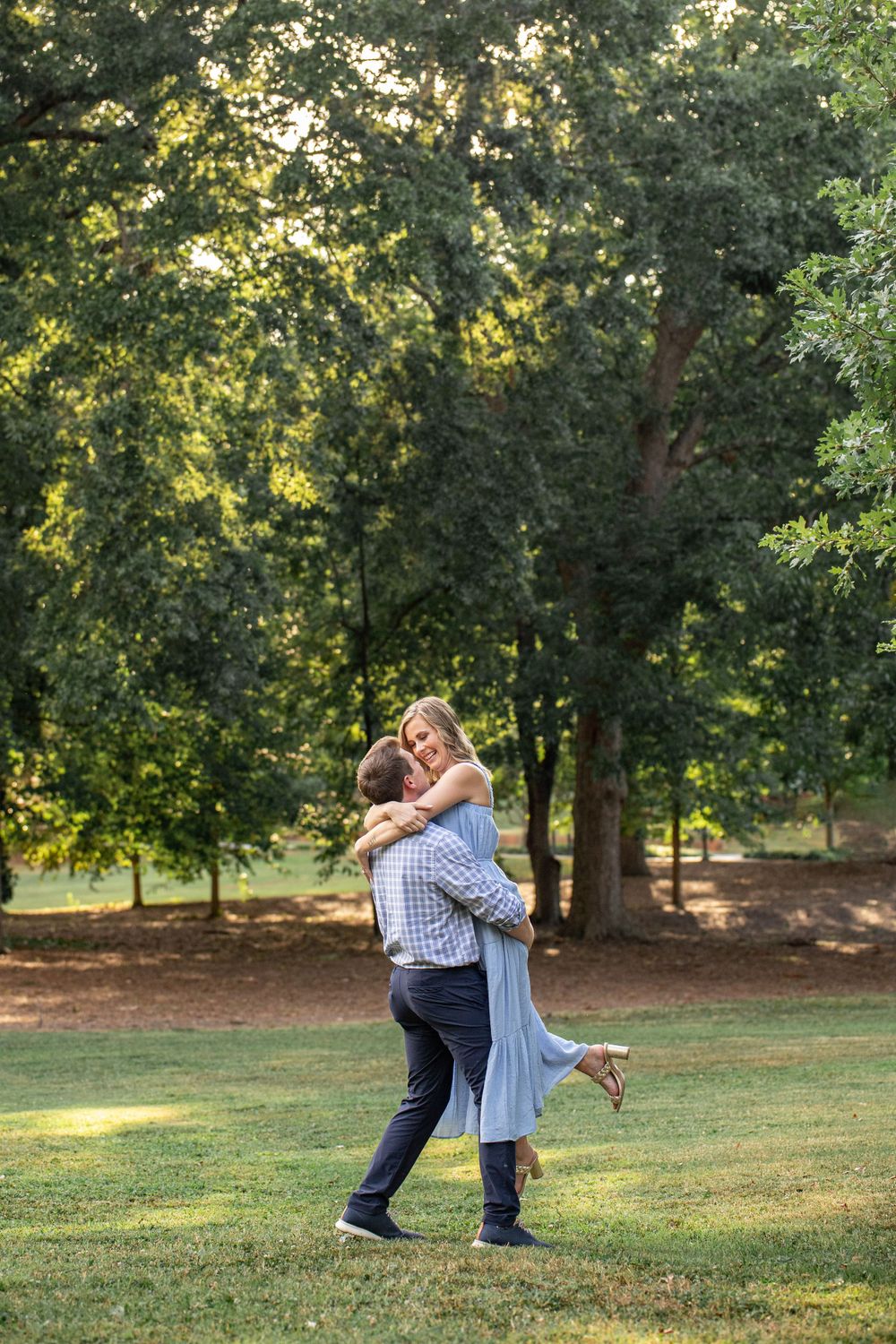 A couple embraces in a romantic moment on a grassy lawn surrounded by sunlit trees in a peaceful park setting.