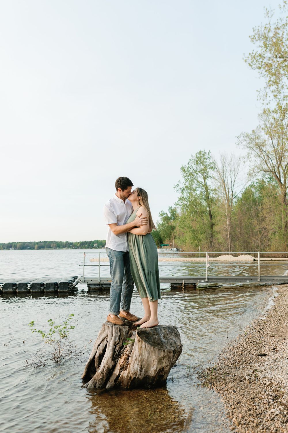 A couple embraces on a large tree stump by a peaceful lakeside.