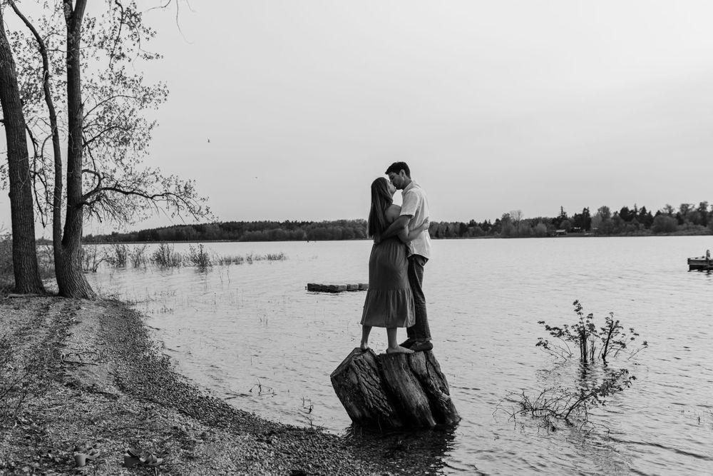 Silhouettes of a couple standing by a serene lake with trees in the background.