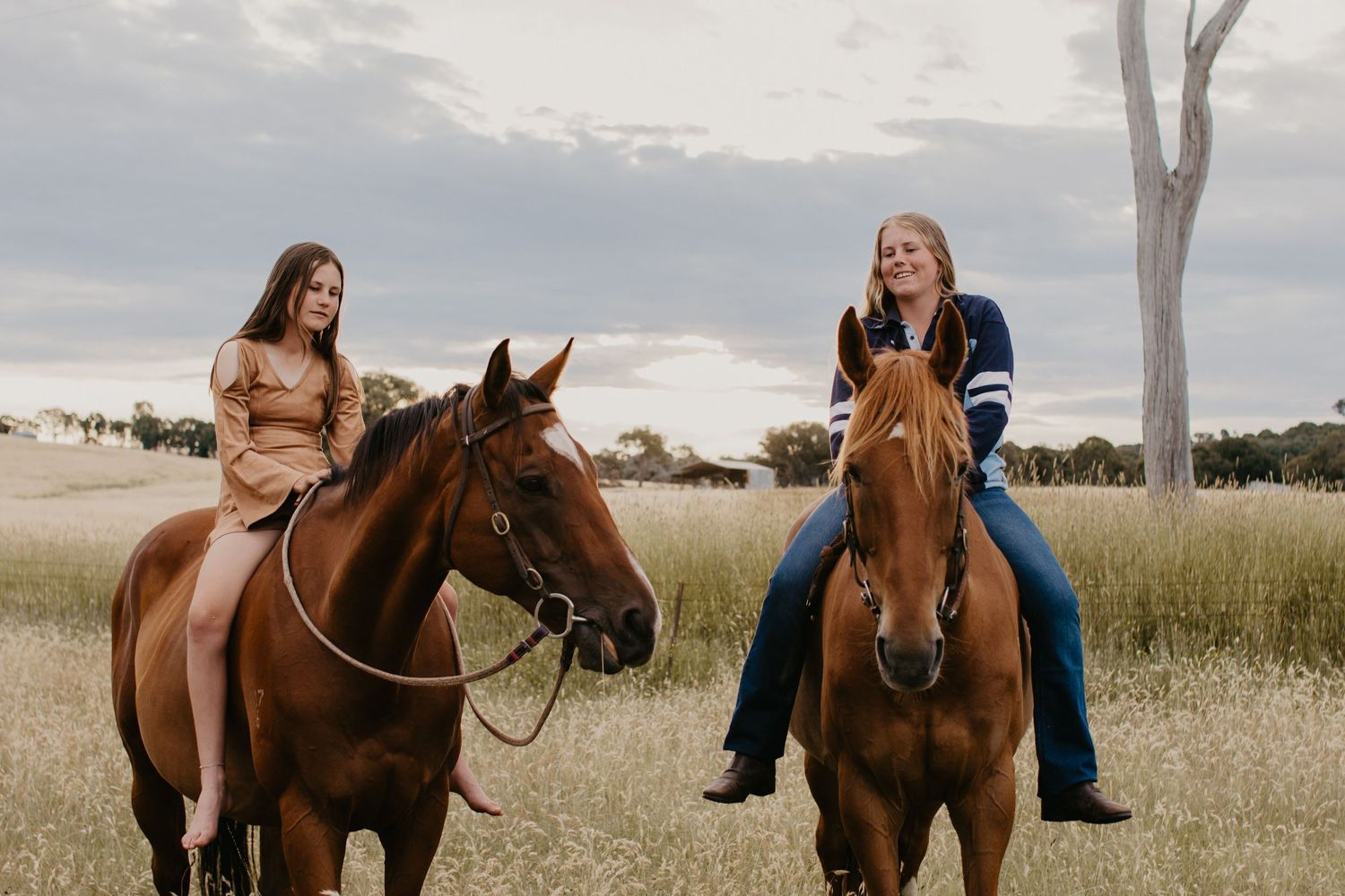 two sister sitting on their horses bareback in the paddock, with a shed and hills in the background on a cloudy day.