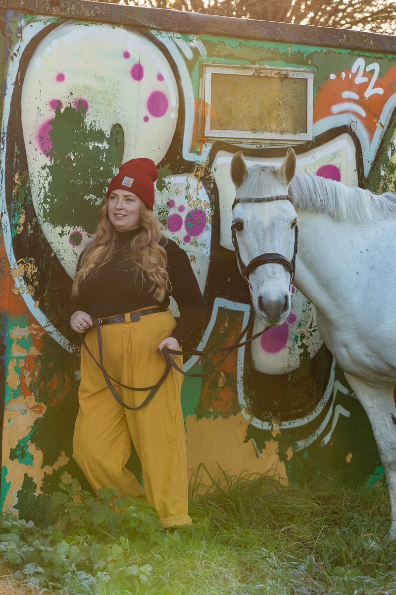 A white horse stands next to street art while someone poses in yellow pants and a red beanie.