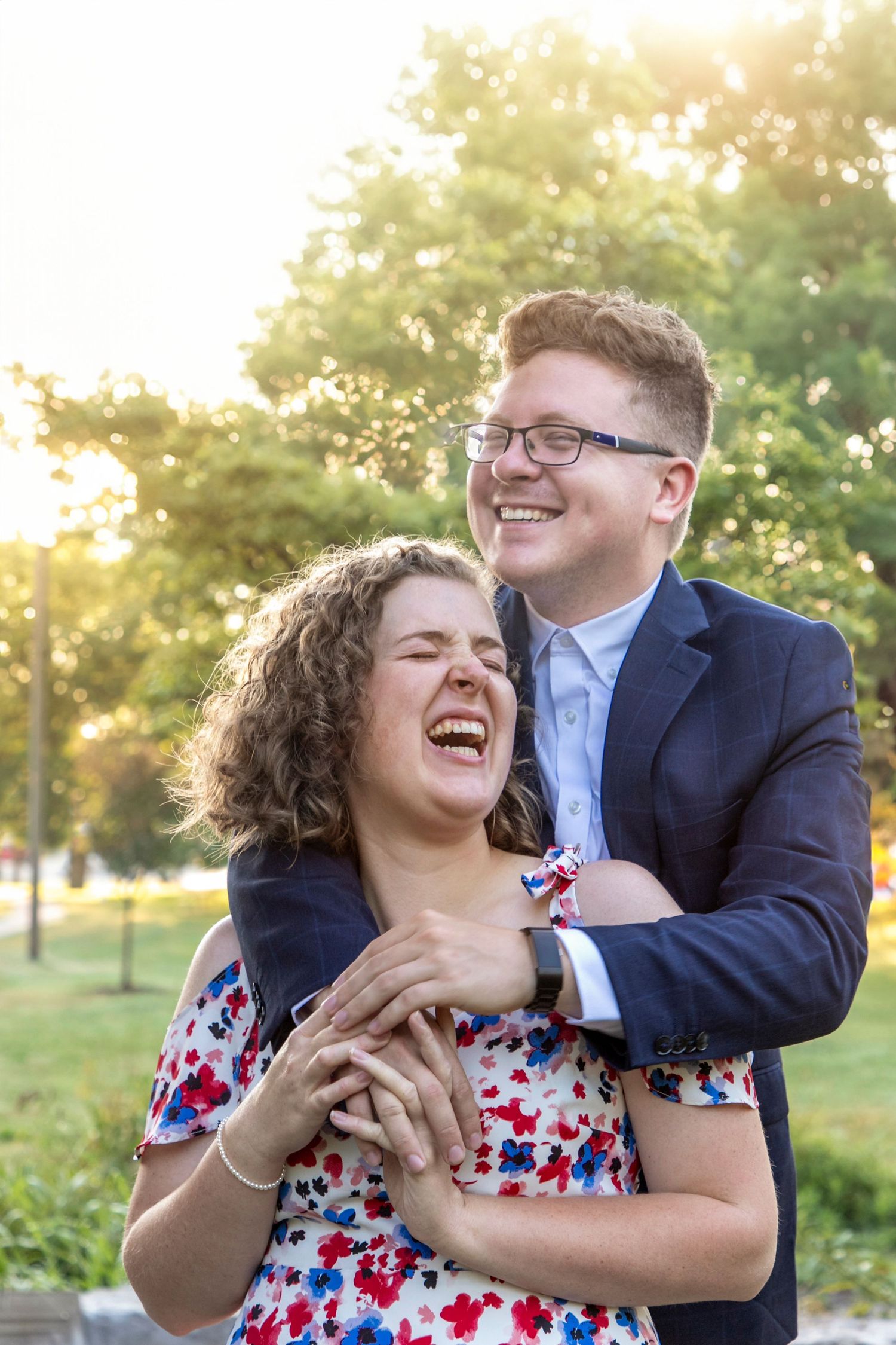 A couple with the guy behind the girl with his arms wrapped around her neck/shoulders looking in opposite directions smiling and laughing