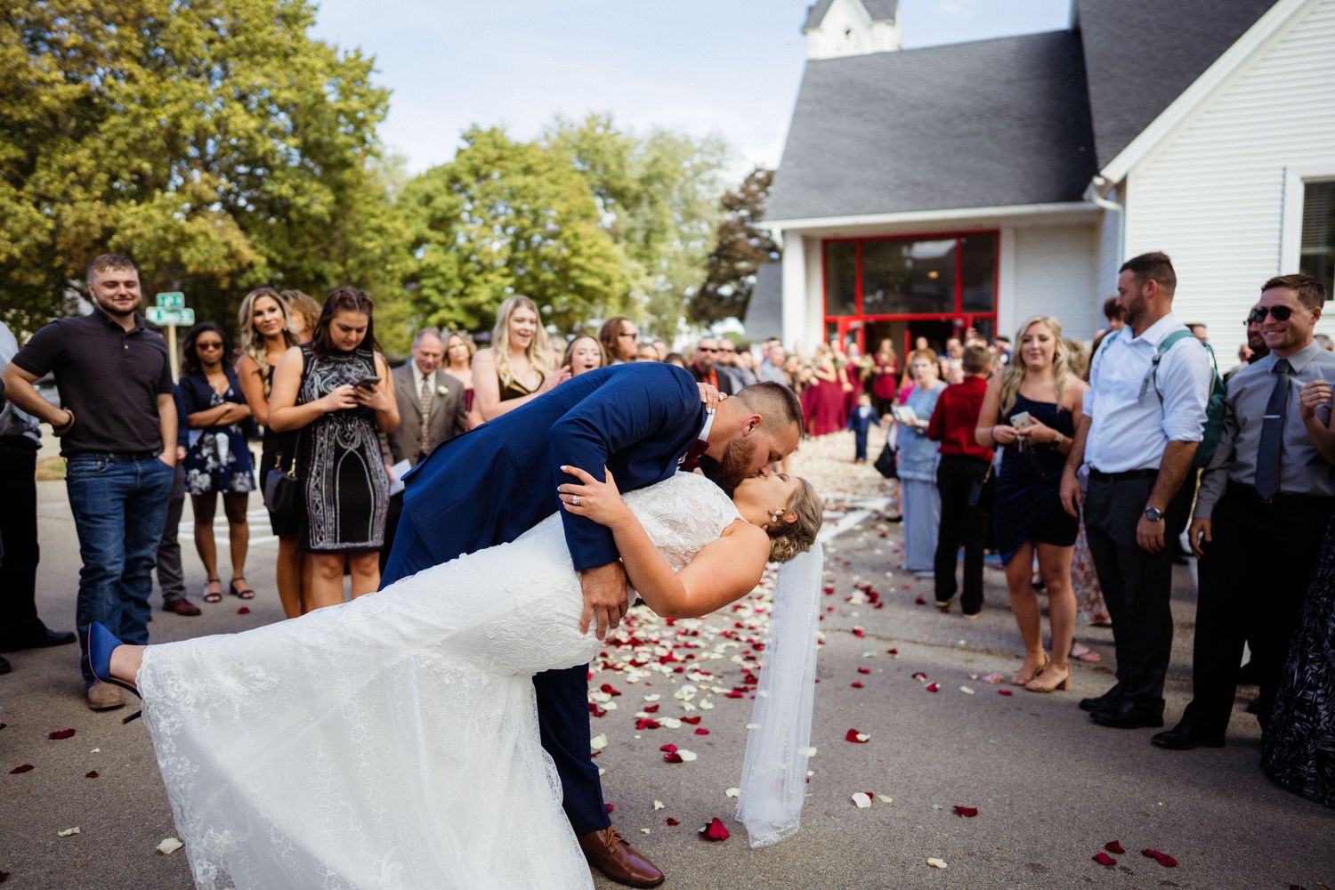 Newlyweds share romantic dip kiss outside church as guests shower them with flower petals.