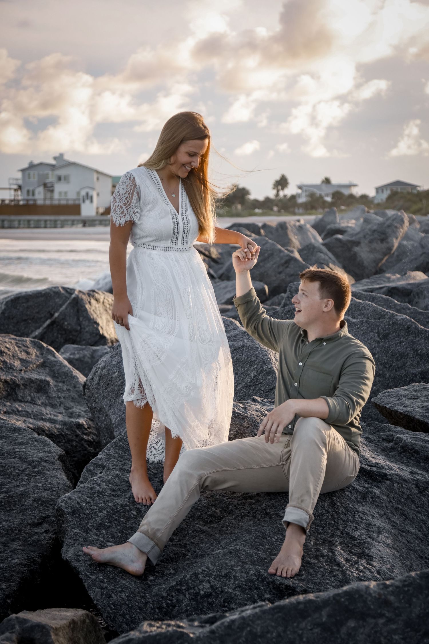 A sunset proposal moment on dark rocky coastline with beach houses in the background.