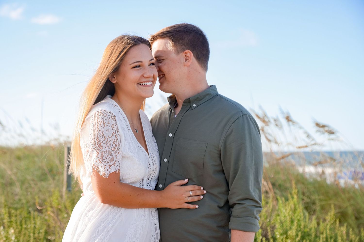 Happy couple embraces in a sunny field with tall grass and mountain views in the background.