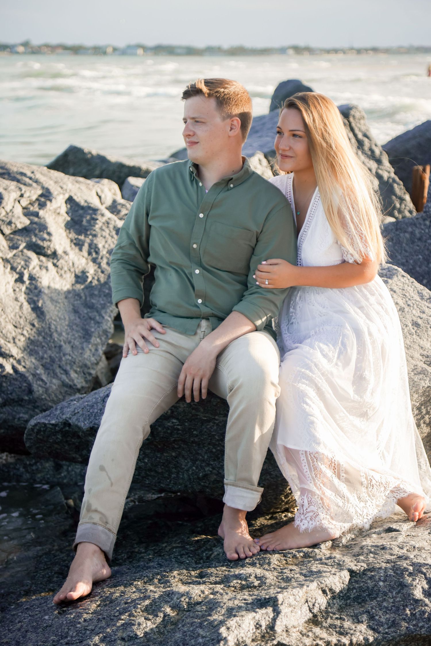 A couple sits barefoot on rocky coastal formations by the ocean during a serene afternoon.