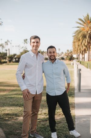 Two men in casual attire pose together on a palm tree-lined pathway.