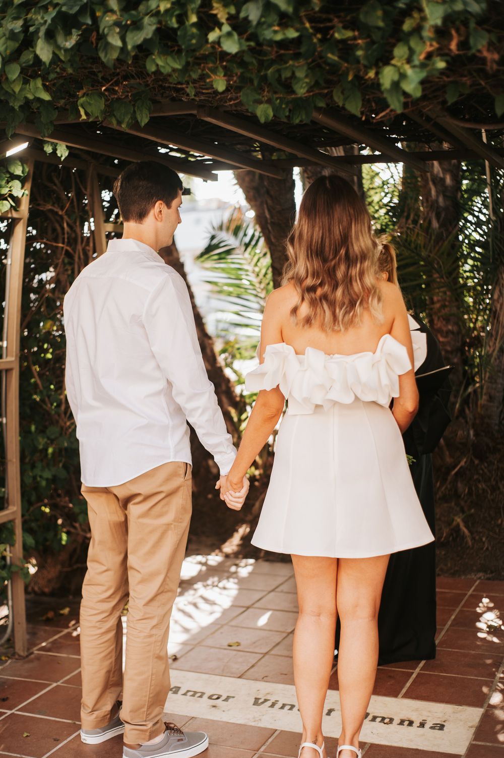 Couple holding hands while walking through a vine-covered pergola.
