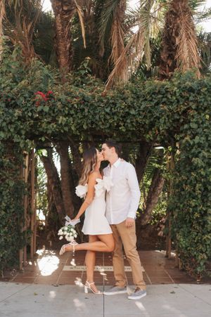 Couple in white and beige outfits share romantic moments in a palm tree garden setting.