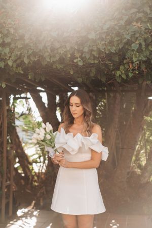 Bride in short white dress standing under vine-covered pergola holding bouquet.