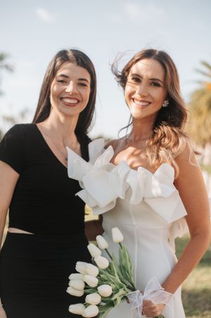 Two women pose together outdoors, one in black and one in white holding white tulips.