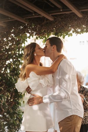 Romantic outdoor wedding kiss under vine-covered pergola with sunlight streaming through.