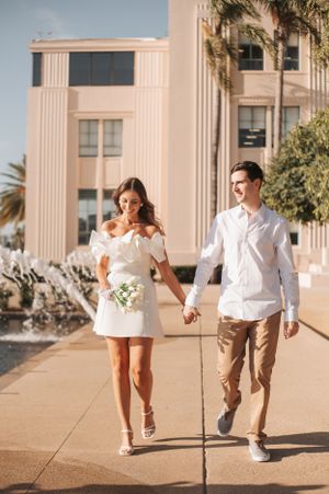 A couple strolls together on a sunny day outside a beige municipal building with palm trees.