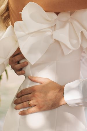 Detail shot of white bow decoration on wedding dress with hands showing wedding ring.