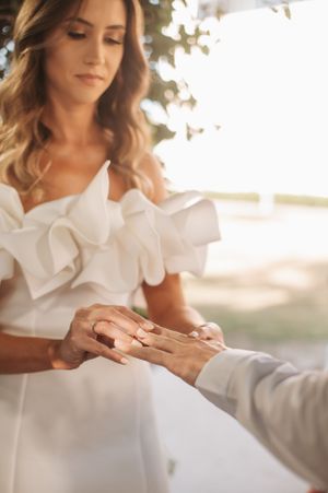 A bride in a white ruffled dress receives a ring during an outdoor wedding ceremony.
