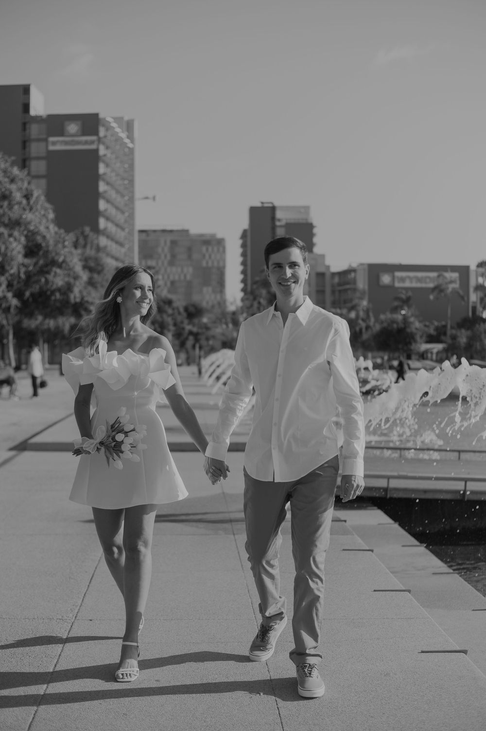 A black and white photo captures a couple walking along an urban pathway with city buildings in background.